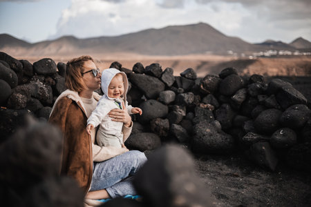 Mother enjoying winter vacations playing with his infant baby boy son on black sandy volcanic beach of Janubio on Lanzarote island, Spain on windy overcast day. Family travel vacations conceptの写真素材