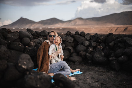 Mother enjoying winter vacations playing with his infant baby boy son on black sandy volcanic beach of Janubio on Lanzarote island, Spain on windy overcast day. Family travel vacations conceptの写真素材
