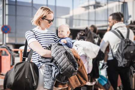 Motherat travelling with his infant baby boy child. Mom holding travel bag and her infant baby boy child while queuing for bus in front of airport terminal stationの写真素材