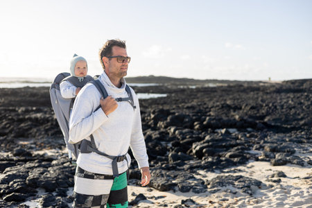 Young father carrying his infant baby boy son in backpack on black rock volcanic beach on Lanzarote island, Spain. Family travel and winter vacation conceptの写真素材