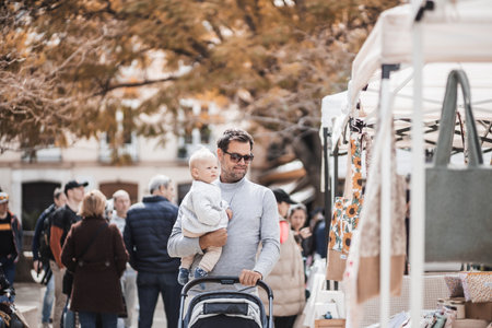 Father walking carrying his infant baby boy child and pushing stroller in crowd of people wisiting sunday flea market in Malaga, Spainの写真素材