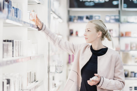 Elegant blond young woman choosing perfume in retail store. Beautiful blond lady testing and buying cosmetics in a beauty store.の写真素材