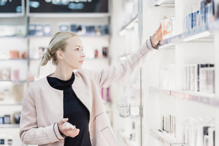 Elegant blond young woman choosing perfume in retail store. Beautiful blond lady testing and buying cosmetics in a beauty store.の写真素材