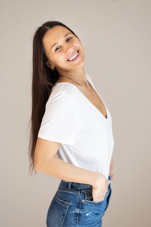 Portrait of confident beautiful woman with long brown hair, wearing casual clothes, standing in relaxed pose with hands in pockets, smiling with white teeth at camera, studio backgroundの写真素材
