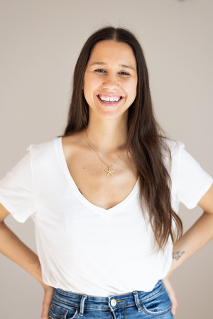 Portrait of confident beautiful woman with long brown hair, wearing casual clothes, standing in relaxed pose with hands in pockets, smiling with white teeth at camera, studio backgroundの写真素材