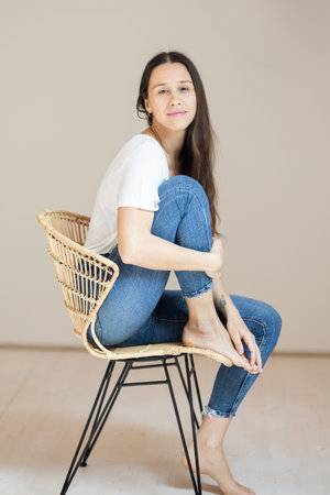 Portrait of confident beautiful woman with long brown hair, wearing casual clothes, sitting on chair in tight jeans and white t-shirt, studio backgroundの写真素材