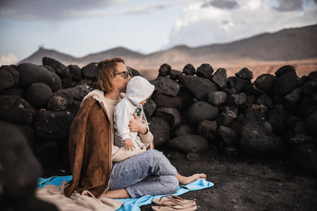 Mother enjoying winter vacations playing with his infant baby boy son on black sandy volcanic beach of Janubio on Lanzarote island, Spain on windy overcast day. Family travel vacations conceptの写真素材