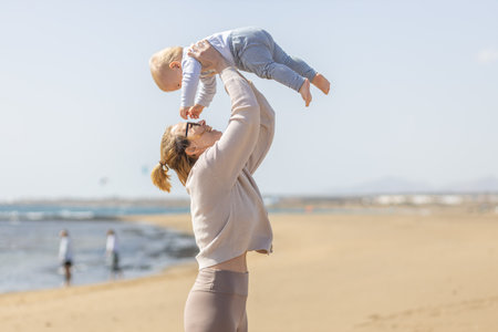 Mother enjoying summer vacations holding, playing and lifting his infant baby boy son high in the air on sandy beach on Lanzarote island, Spain. Family travel and vacations conceptの写真素材