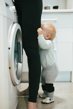 Little infant baby boy child hiding between mothers legs demanding her attention while she is multitasking, trying to do some household chores in kitchen at home. Mother on maternity leaveの写真素材