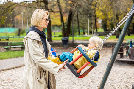 Mother pushing her infant baby boy child wearing yellow rain boots and cape on swing on playground outdoors on cold rainy overcast autumn day in Ljubljana, Slovenia.の写真素材