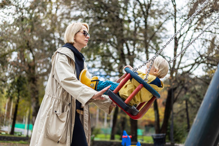 Mother pushing her infant baby boy child wearing yellow rain boots and cape on swing on playground outdoors on cold rainy overcast autumn day in Ljubljana, Slovenia.の写真素材