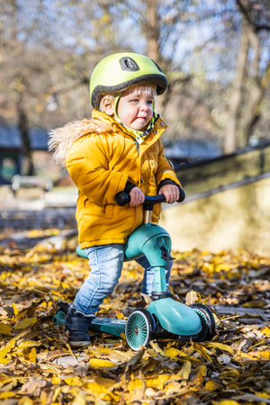Adorable toddler boy wearing yellow protective helmet riding baby scooter outdoors on autumn day. Kid training balance on mini bike in city park. Fun autumn outdoor activity for small kidsの写真素材
