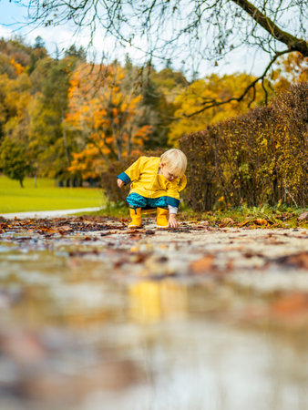 Sun always shines after the rain. Small bond infant boy wearing yellow rubber boots and yellow waterproof raincoat walking in puddles in city park on sunny rainy dayの写真素材