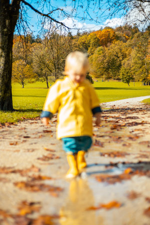 Sun always shines after the rain. Small bond infant boy wearing yellow rubber boots and yellow waterproof raincoat walking in puddles in city park on sunny rainy dayの写真素材