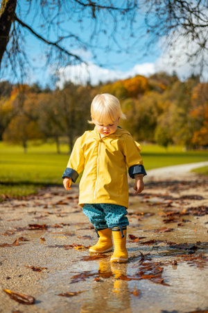 Sun always shines after the rain. Small bond infant boy wearing yellow rubber boots and yellow waterproof raincoat walking in puddles in city park on sunny rainy dayの写真素材