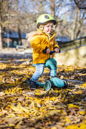 Adorable toddler boy wearing yellow protective helmet riding baby scooter outdoors on autumn day. Kid training balance on mini bike in city park. Fun autumn outdoor activity for small kidsの写真素材