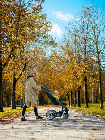Young beautiful mother wearing a rain coat pushing stroller with her little baby boy child, walking in city park on a sunny autumn day.の写真素材