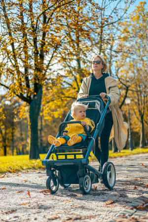 Young beautiful mother wearing a rain coat pushing stroller with her little baby boy child, walking in city park on a sunny autumn day.の写真素材