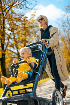 Young beautiful mother wearing a rain coat pushing stroller with her little baby boy child, walking in city park on a sunny autumn day.の写真素材