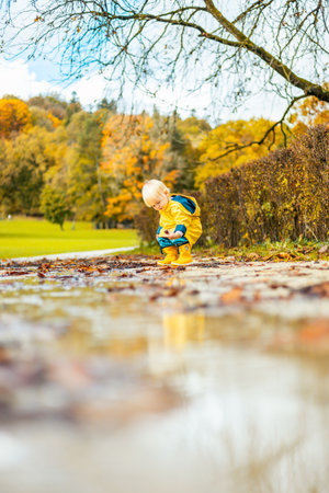 Sun always shines after the rain. Small bond infant boy wearing yellow rubber boots and yellow waterproof raincoat walking in puddles in city park on sunny rainy dayの写真素材