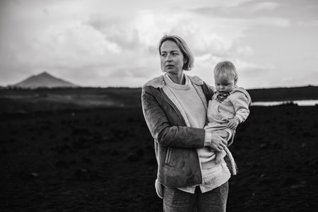 Mother enjoying winter vacations playing with his infant baby boy son on black sandy volcanic beach of Janubio on Lanzarote island, Spain on windy overcast day. Family travel vacations conceptの写真素材