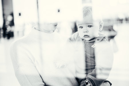 Thoughtful young mother looking trough window holding his infant baby boy child while waiting to board an airplane at airport terminal departure gates. Travel with baby conceptの写真素材