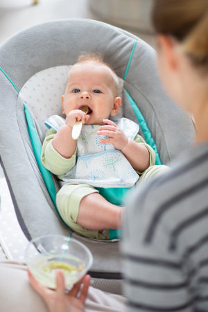 Mother spoon feeding her baby boy child in baby chair with fruit puree. Baby solid food introduction conceptの写真素材
