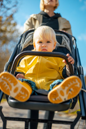 Little toddler child wearing yellow rain coat and rain boots being pushed in stroller by her mother in city park on a sunny autumn day after the rains were goneの写真素材
