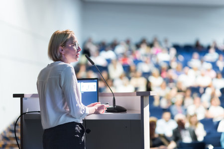 Female speaker giving a talk on corporate business conference. Unrecognizable people in audience at...