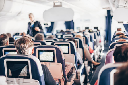 Interior of airplane with passengers on seats and stewardess in uniform walking the aisle, serving...