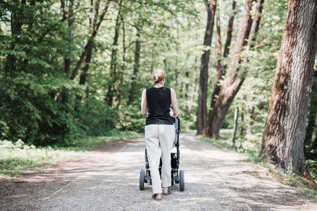 Rear view of casually dressed mother walking with baby stroller in Koseski bajer city park in Ljubljana, Slovenia. Family, child and parenthood concept.の写真素材