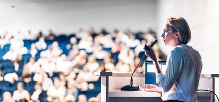 Female speaker giving a talk on corporate business conference. Unrecognizable people in audience at...