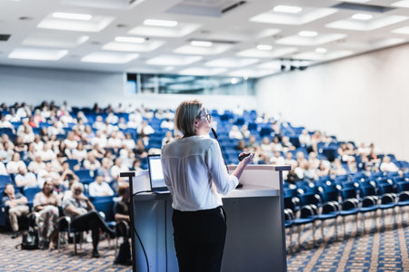Female speaker giving a talk on corporate business conference. Unrecognizable people in audience at...