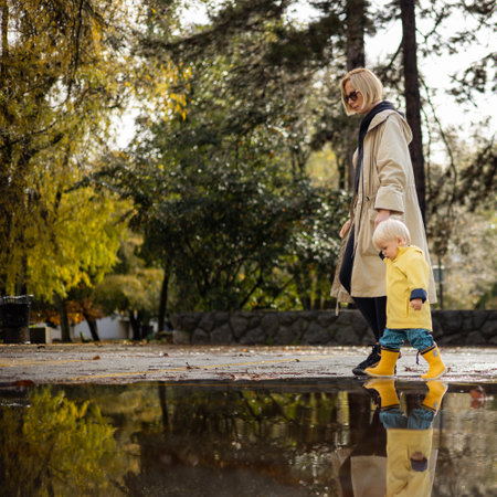 Small bond infant boy wearing yellow rubber boots and yellow waterproof raincoat walking in puddles on a overcast rainy day holding her mothers hand. Mom with small child in rain outdoorsの写真素材
