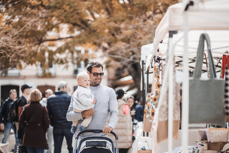 Father walking carrying his infant baby boy child and pushing stroller in crowd of people visiting Sunday flea market in Malaga, Spainの写真素材