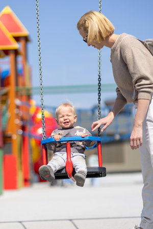 Mother pushing her infant baby boy child on a swing on playground outdoorsの写真素材