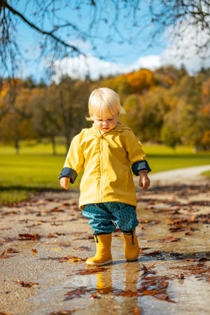 Sun always shines after the rain. Small bond infant boy wearing yellow rubber boots and yellow waterproof raincoat walking in puddles in city park on sunny rainy dayの写真素材