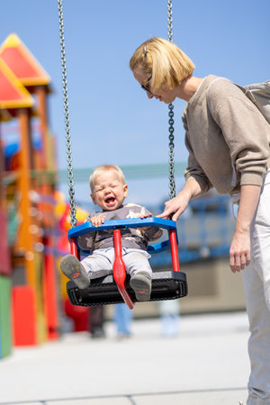 Mother pushing her infant baby boy child on a swing on playground outdoorsの写真素材