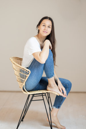Portrait of confident beautiful woman with long brown hair, wearing casual clothes, sitting on chair in tight jeans and white t-shirt, studio backgroundの写真素材