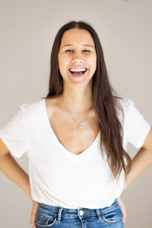 Portrait of confident beautiful woman with long brown hair, wearing casual clothes, standing in relaxed pose with hands in pockets, smiling with white teeth at camera, studio backgroundの写真素材