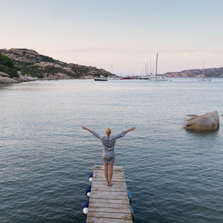 Beautiful woman standing on wooden pier arms rised enjoying peaceful seascape at dusk. Female traveler stands on a wooden pier in Porto Rafael, Costa Smeralda, Sardinia, Italy.の写真素材