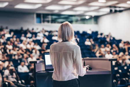Female speaker giving a talk on corporate business conference. Unrecognizable people in audience at conference hall. Business and Entrepreneurship eventの写真素材
