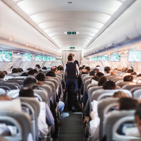 Interior of airplane with passengers on seats and stewardess in uniform walking the aisle, serving...