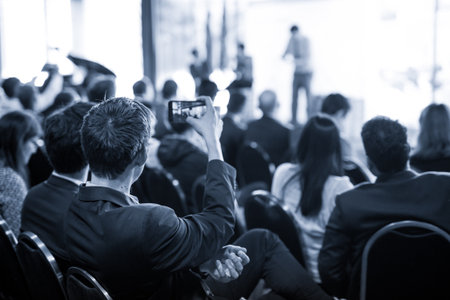 Businessman takes a picture of corporate business presentation at conference hall using smartphone....
