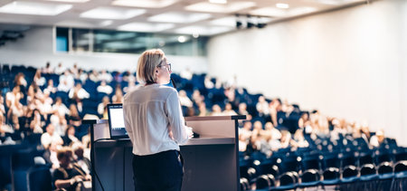 Female speaker giving a talk on corporate business conference. Unrecognizable people in audience at...