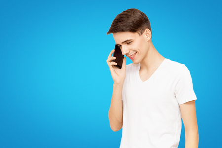 a guy in a white T-shirt talking on the phone on a blue isolated background, talkative young man, joyful man in lifeの写真素材