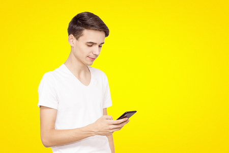 The guy in the white T-shirt with the phone on a yellow background. Young teenager prescribed in social networks, the concept of modern technologyの写真素材