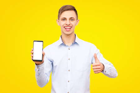 guy in a blue shirt advertises a phone isolated on a yellow background in the studio, showing thumb upの写真素材