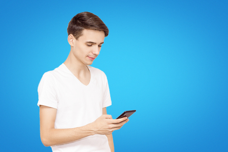 The guy in the white T-shirt with the phone on a blue background. Young teenager prescribed in social networks, the concept of modern technologyの写真素材