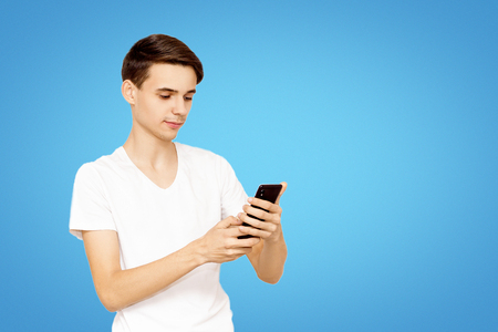 The guy in the white T-shirt with the phone on a blue background. Young teenager prescribed in social networks, the concept of modern technologyの写真素材